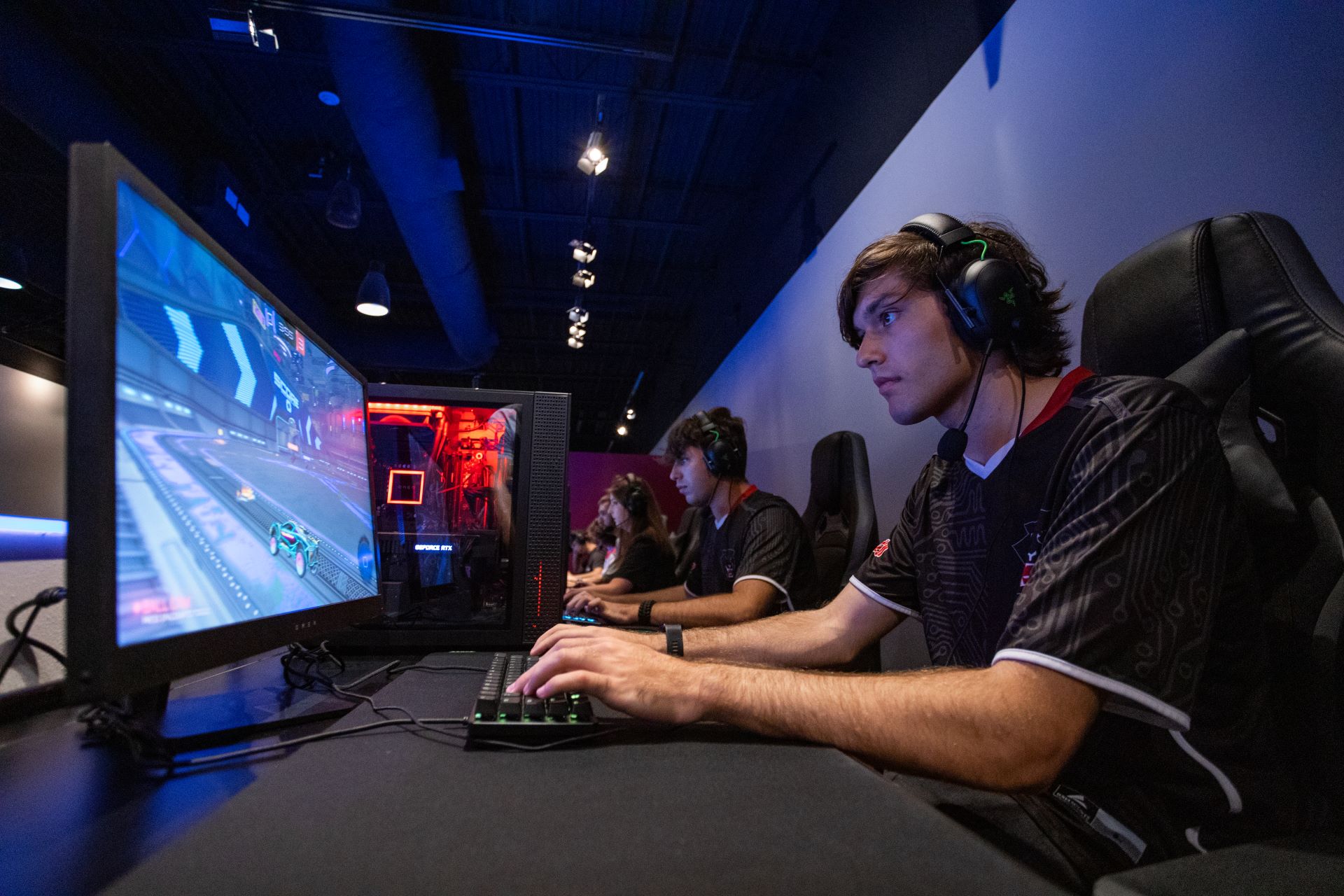 Close-up view of a student intensely focused on playing a game on a computer at the Esports center, wearing a headset and gaming attire, with other students engaged in gaming in the background